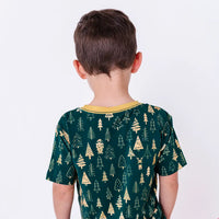 Back view of a child in a short sleeve tee with a mustard ribbed collar in a dark green print of gold Christmas trees.