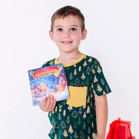 A child smiling at the camera while holding a storybook, in a short sleeve pocket tee with a mustard pocket in a dark green print of gold Christmas trees.