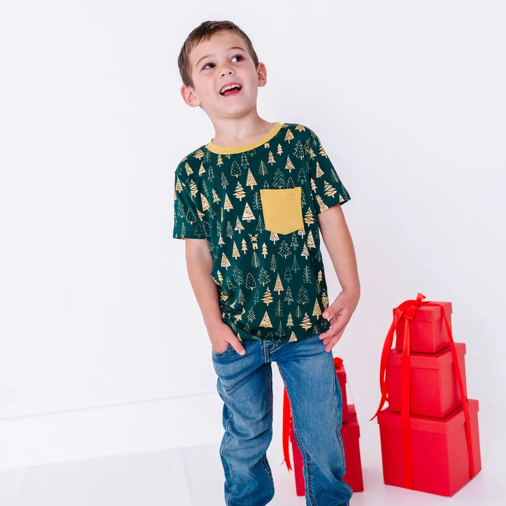 A child laughing and looking up to the side with both hands in the jeans pockets, in a short sleeve pocket tee in a dark green print of gold Christmas trees.