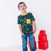 A child laughing and looking up to the side with both hands in the jeans pockets, in a short sleeve pocket tee in a dark green print of gold Christmas trees.