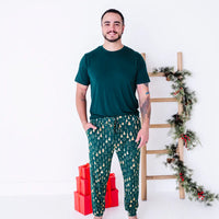 A man smiling and standing with hands in the pockets, wearing a dark green tee and jogger pajama pants in a dark green print of gold Christmas trees.