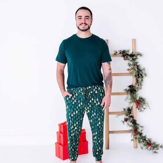 A man smiling and standing with hands in the pockets, wearing a dark green tee and jogger pajama pants in a dark green print of gold Christmas trees.