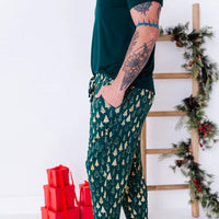 Side view of a man with a hand in the pocket of jogger pajama pants in a dark green print of gold Christmas trees.