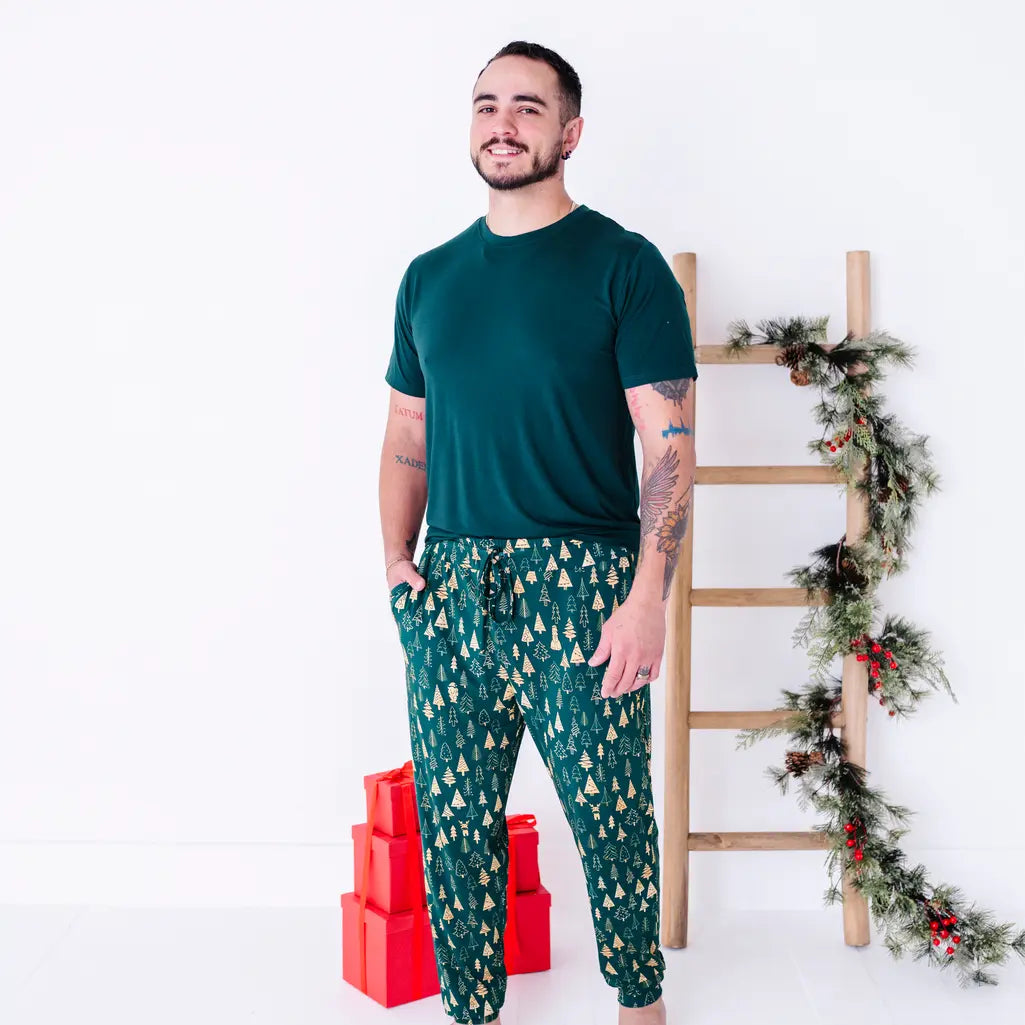 A man smiling and turned slightly to one side with a hand in the pocket, in a dark green tee and jogger pajama pants in a dark green print of gold Christmas trees.