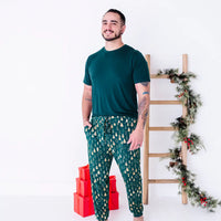 A man smiling and turned slightly to one side with a hand in the pocket, in a dark green tee and jogger pajama pants in a dark green print of gold Christmas trees.