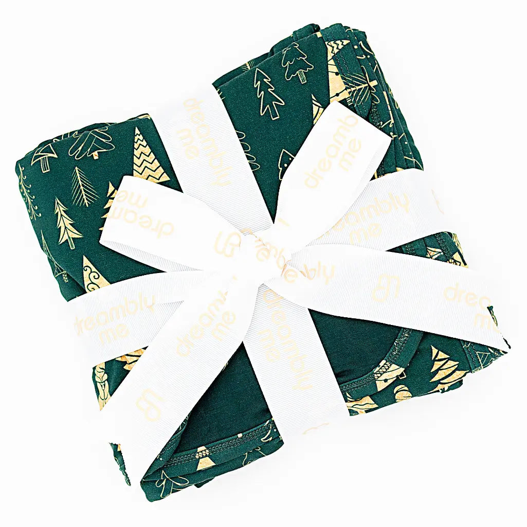 Flat lay of a folded dark green blanket tied with a white ribbon in a print of gold and outlined Christmas trees on a white background.