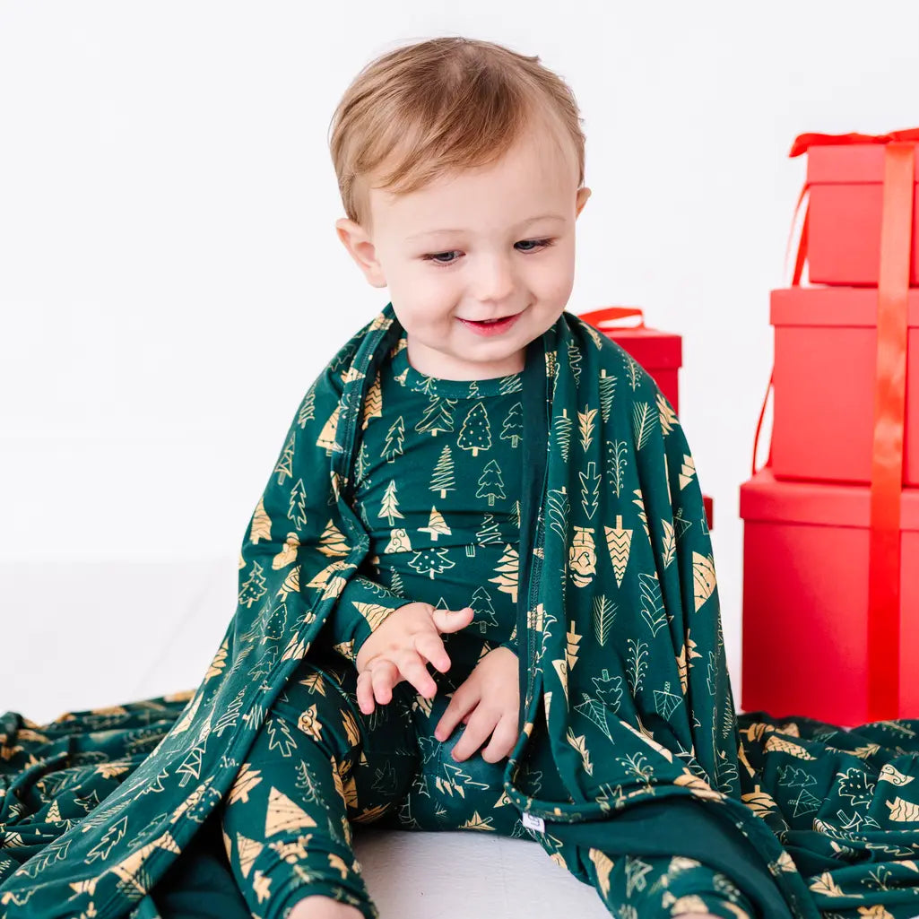 A smiling toddler in matching pajamas sitting on the floor with a dark green blanket in a print of gold and outlined Christmas trees draped over the shoulders and spread around.