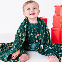 A toddler looking up with a big smile, a dark green blanket in a print of gold and outlined Christmas trees draped around the shoulders, with red gift boxes behind.