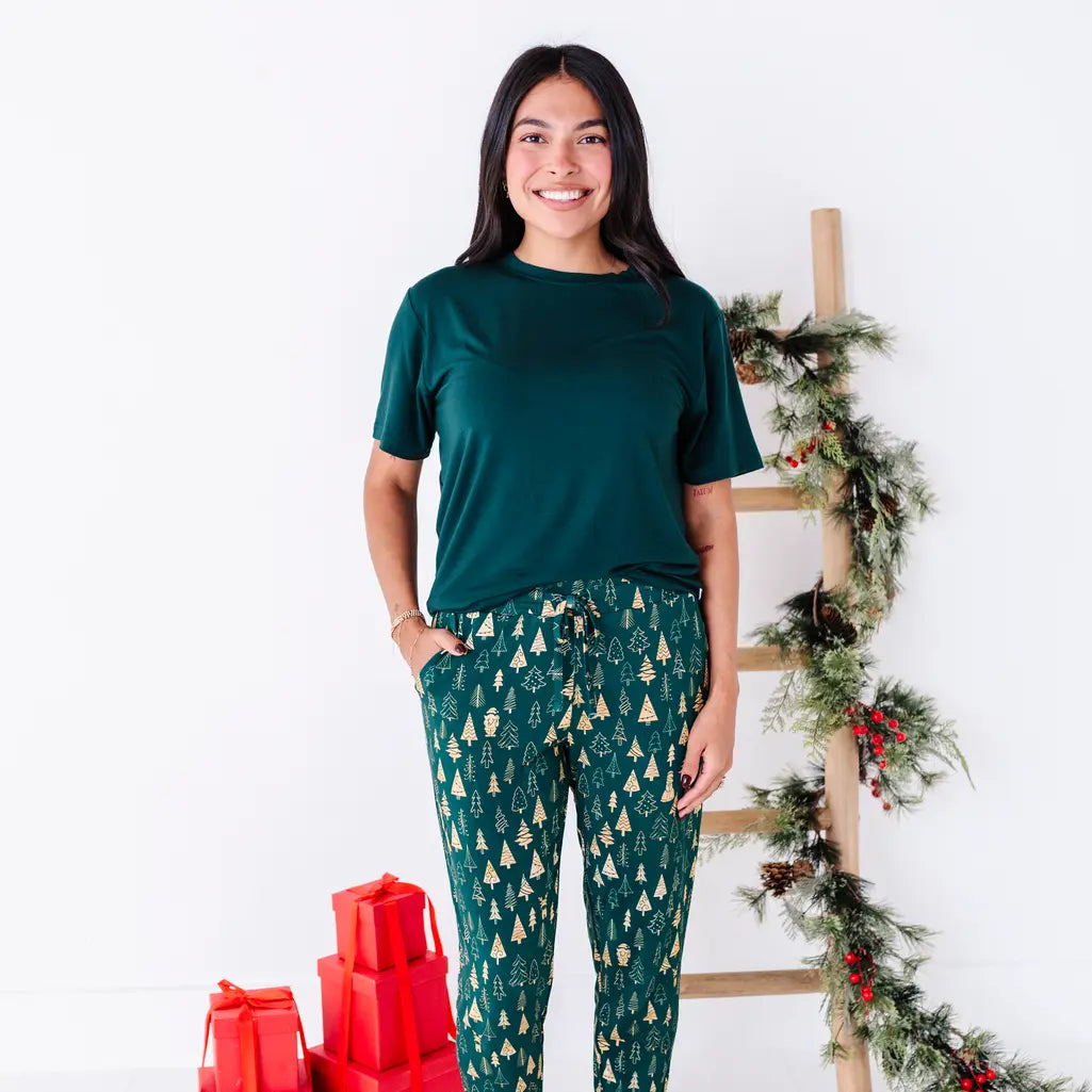 A woman with a big smile standing with hands in the pockets, wearing a dark green tee and jogger pajama pants in a dark green print of gold Christmas trees.