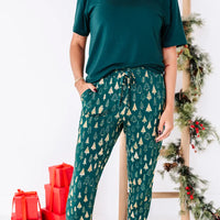 Close up of the waist and legs of jogger pajama pants with a drawstring and pocket in a dark green print of gold Christmas trees.