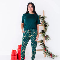 A woman smiling with one leg forward and a hand in the pocket, in a dark green tee and jogger pajama pants in a dark green print of gold Christmas trees.