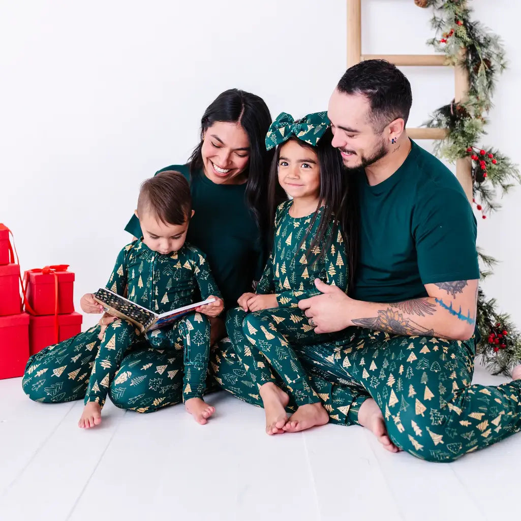 A family of four sitting on the floor reading a book together in matching pajamas in a dark green print of gold Christmas trees and dark green tees.