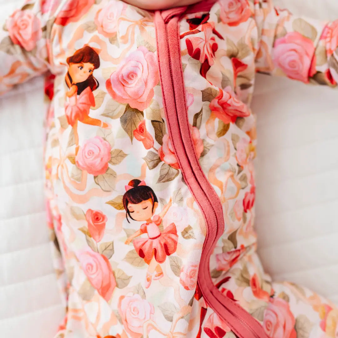Close-up of a zip-up baby pajama with a print of pink roses, ribbons, and little ballerinas, with rose-pink zipper trim.