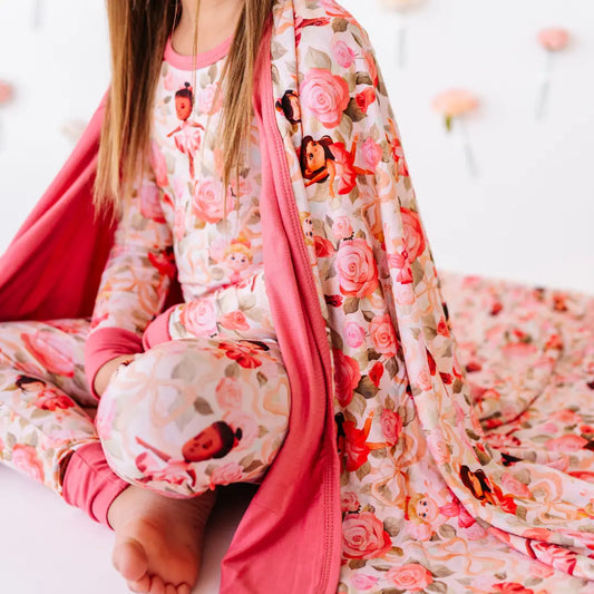 A child sitting cross-legged in rose and ballerina print pajamas draped in a matching blanket with a rose-pink underside.