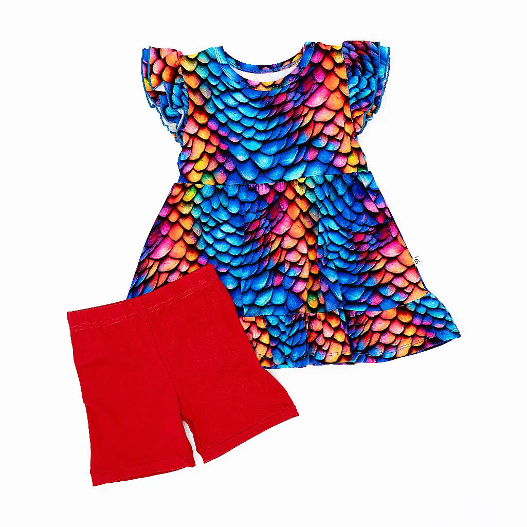 Flat lay of a flutter sleeve peplum top in a rainbow dragon scale print with red bike shorts on a white background.