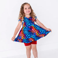 A child with curly hair smiling and holding out the hem of a flutter sleeve peplum top in a rainbow dragon scale print with red bike shorts.