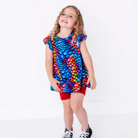 A child with curly hair looking up and smiling, in a flutter sleeve peplum top in a rainbow dragon scale print with red bike shorts.
