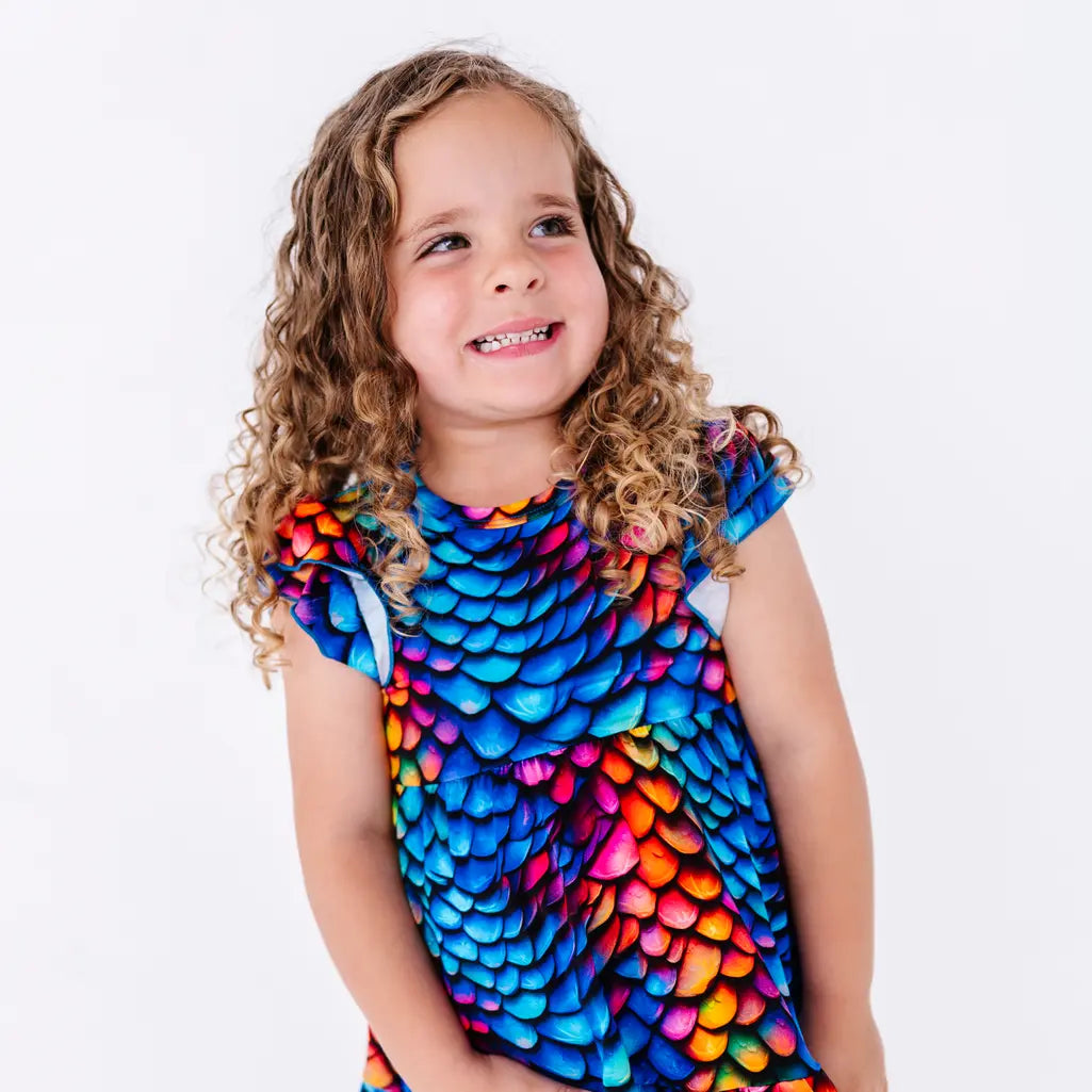 Close up portrait of a child with curly hair smiling with the head tilted, in a flutter sleeve peplum top in a rainbow dragon scale print.