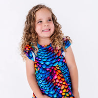 Close up portrait of a child with curly hair smiling with the head tilted, in a flutter sleeve peplum top in a rainbow dragon scale print.