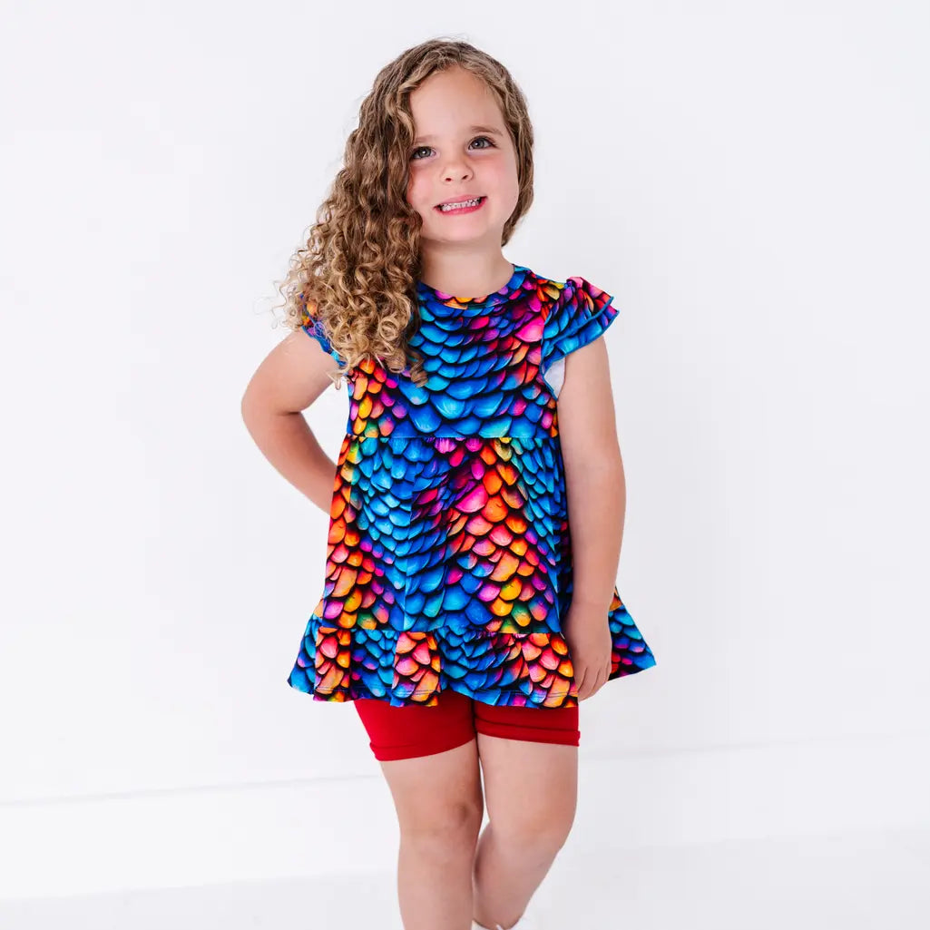 A child with curly hair standing with one hand on the hip, in a flutter sleeve peplum top in a rainbow dragon scale print with red bike shorts.