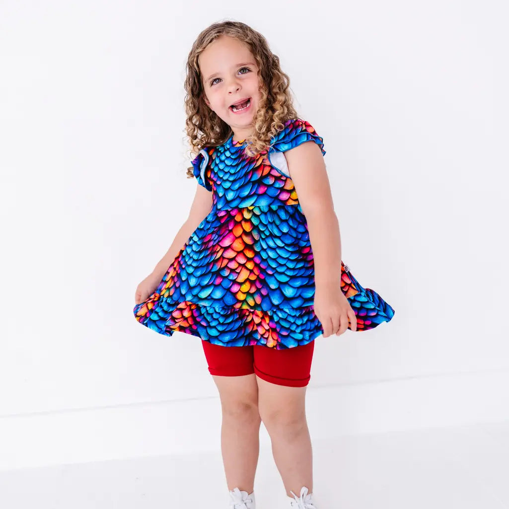 A child laughing and swishing the hem of a flutter sleeve peplum top in a rainbow dragon scale print with red bike shorts.