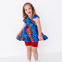 A child laughing and swishing the hem of a flutter sleeve peplum top in a rainbow dragon scale print with red bike shorts.
