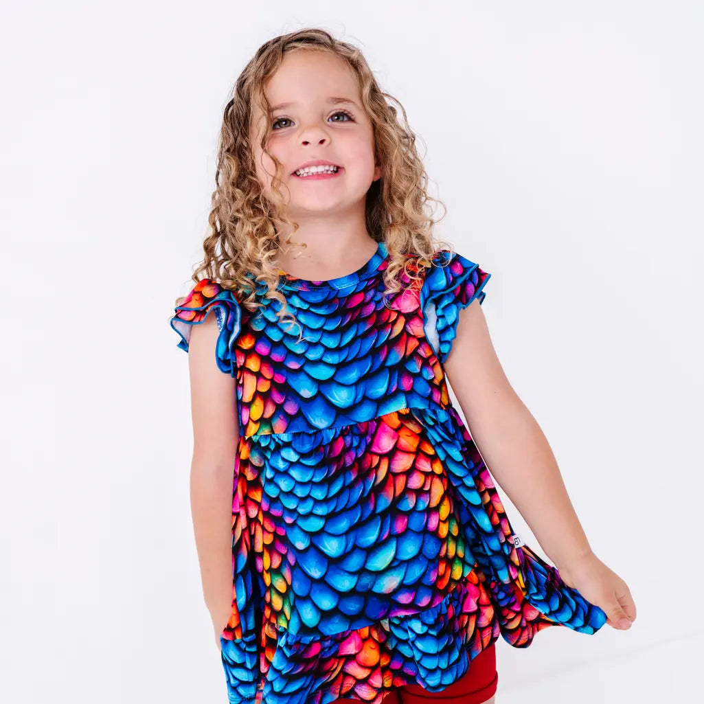 A child with curly hair smiling at the camera in a flutter sleeve peplum top in a rainbow dragon scale print, holding out the hem.