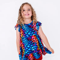 A child with curly hair smiling at the camera in a flutter sleeve peplum top in a rainbow dragon scale print, holding out the hem.
