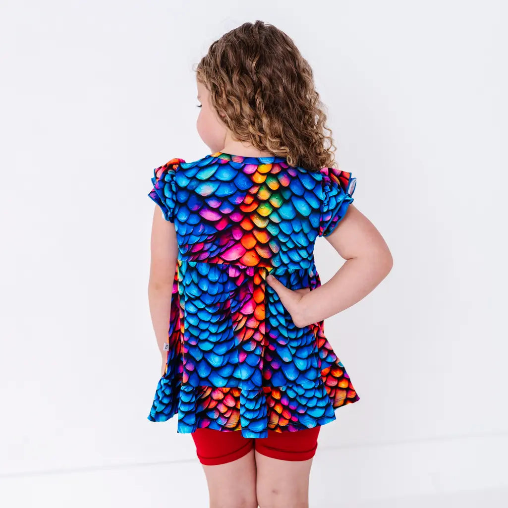 Back view of a child with a hand on the hip and the head turned, in a flutter sleeve peplum top in a rainbow dragon scale print with red bike shorts.