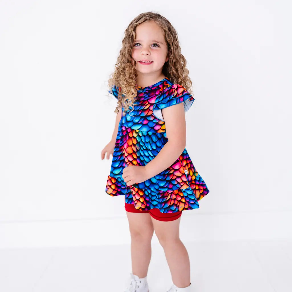 A child in a three quarter pose smiling at the camera, in a flutter sleeve peplum top in a rainbow dragon scale print with red bike shorts.