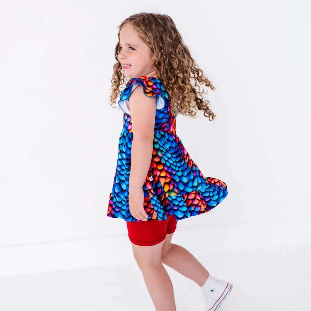 Side view of a child mid spin with swinging curly hair, in a flutter sleeve peplum top in a rainbow dragon scale print with red bike shorts.