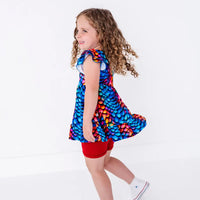 Side view of a child mid spin with swinging curly hair, in a flutter sleeve peplum top in a rainbow dragon scale print with red bike shorts.