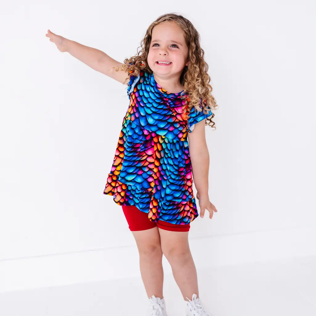 A child smiling with one arm stretched out to the side, in a flutter sleeve peplum top in a rainbow dragon scale print with red bike shorts.
