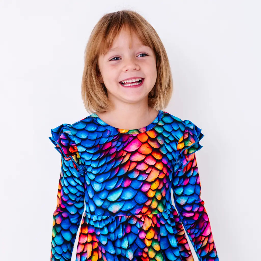 Close up portrait of a child with a big open smile, in a long sleeve twirl dress with flutter ruffles at the shoulders in a rainbow dragon scale print of blue, pink, orange, and yellow.