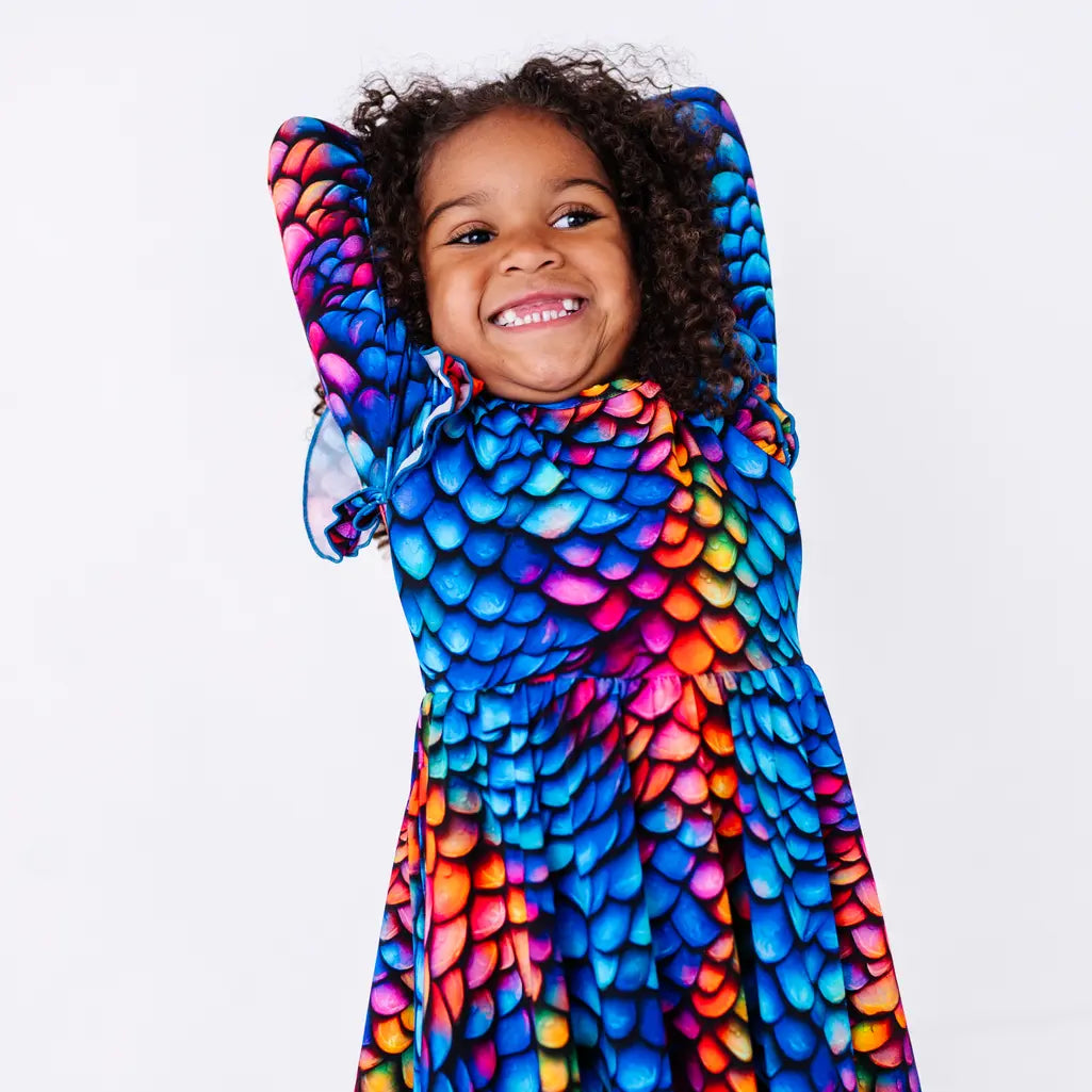 A grinning child with an arm raised behind the head, in a long sleeve twirl dress with flutter ruffles at the shoulders in a rainbow dragon scale print of blue, pink, orange, and yellow.