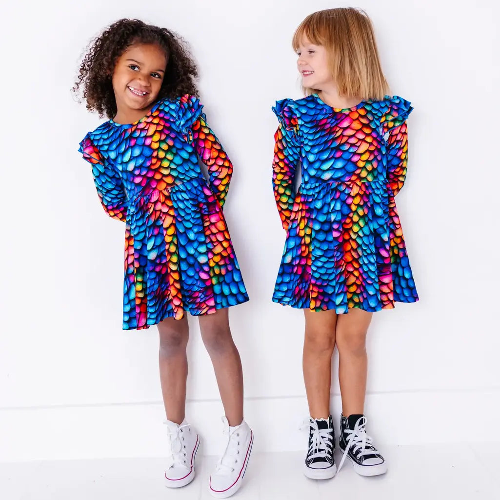 Two kids in matching long sleeve twirl dresses, one beaming at the camera, in a rainbow dragon scale print of blue, pink, orange, and yellow.