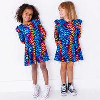 Two kids in matching long sleeve twirl dresses, one beaming at the camera, in a rainbow dragon scale print of blue, pink, orange, and yellow.
