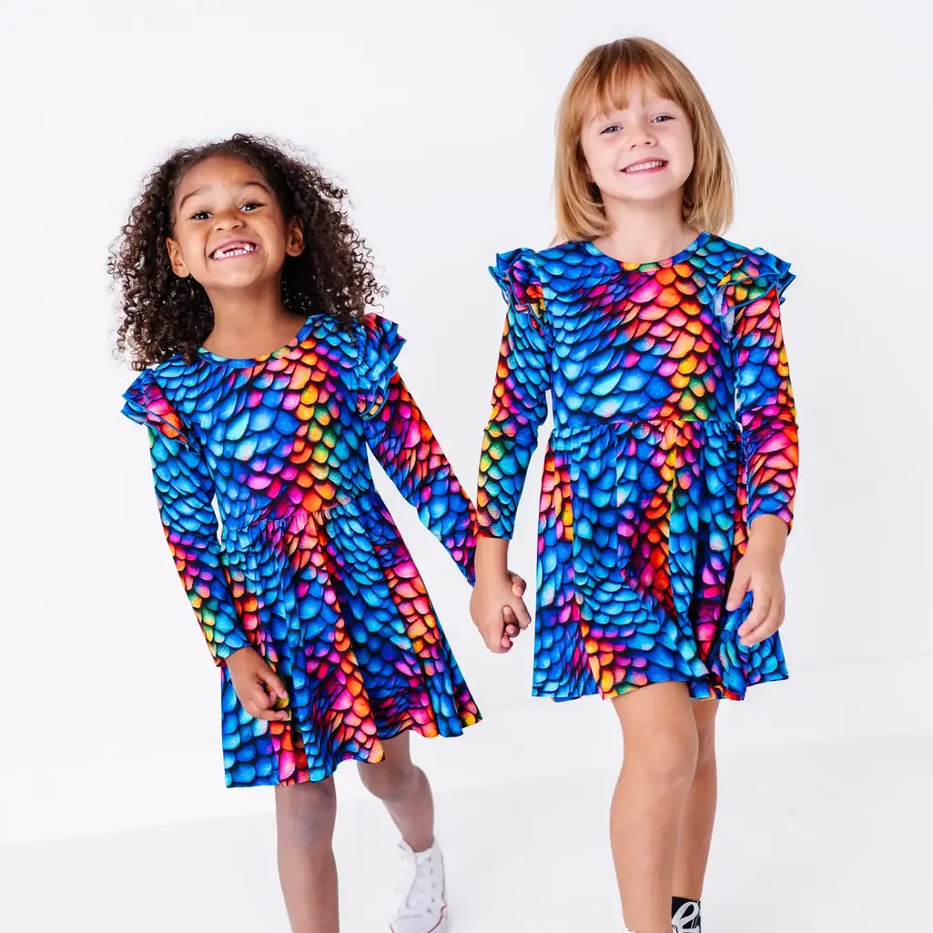 Two kids walking hand in hand and grinning in matching long sleeve twirl dresses in a rainbow dragon scale print of blue, pink, orange, and yellow.