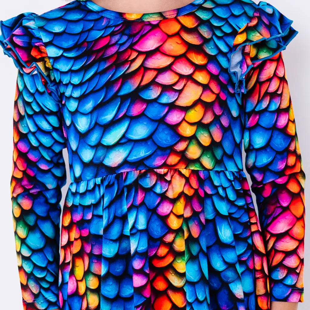 Close up of the front of a long sleeve twirl dress with flutter ruffles at the shoulders in a rainbow dragon scale print of blue, pink, orange, and yellow.