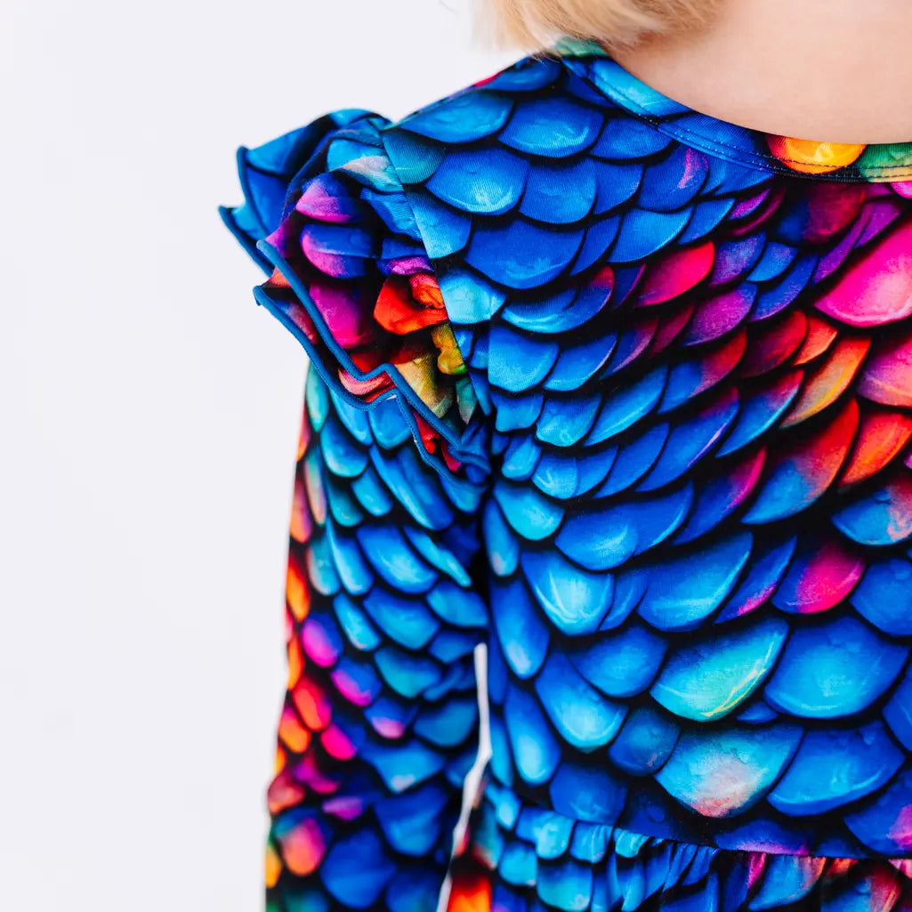 Close up of a flutter ruffle on the shoulder of a long sleeve twirl dress in a rainbow dragon scale print of blue, pink, orange, and yellow.