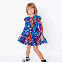 A child smiling and walking with the skirt swinging, in a long sleeve twirl dress with flutter ruffles at the shoulders in a rainbow dragon scale print of blue, pink, orange, and yellow.