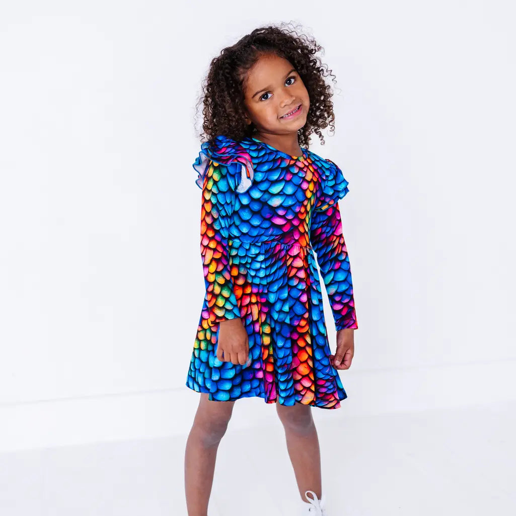 A child with curly hair smiling gently with the body turned, in a long sleeve twirl dress with flutter ruffles at the shoulders in a rainbow dragon scale print of blue, pink, orange, and yellow.