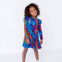 A child with curly hair smiling gently with the body turned, in a long sleeve twirl dress with flutter ruffles at the shoulders in a rainbow dragon scale print of blue, pink, orange, and yellow.
