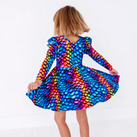 Back view of a child twirling with the head turned, in a long sleeve twirl dress with flutter ruffles at the shoulders in a rainbow dragon scale print of blue, pink, orange, and yellow.