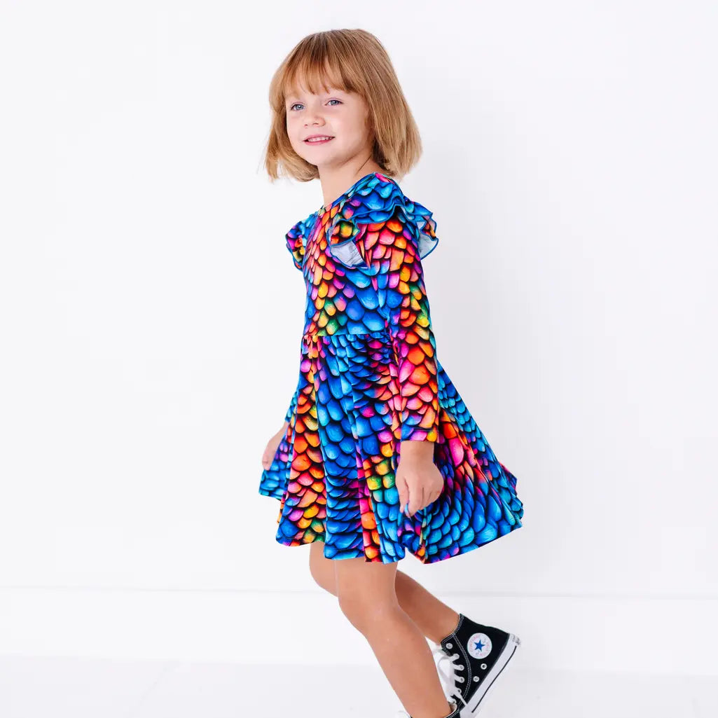 Side view of a smiling child mid step, in a long sleeve twirl dress with flutter ruffles at the shoulders in a rainbow dragon scale print of blue, pink, orange, and yellow.