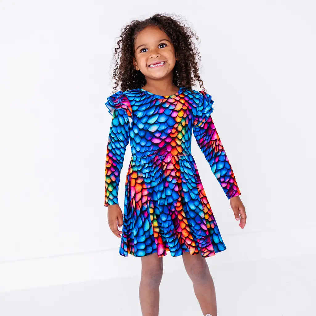A child with curly hair walking with arms out and a smile, in a long sleeve twirl dress with flutter ruffles at the shoulders in a rainbow dragon scale print of blue, pink, orange, and yellow.