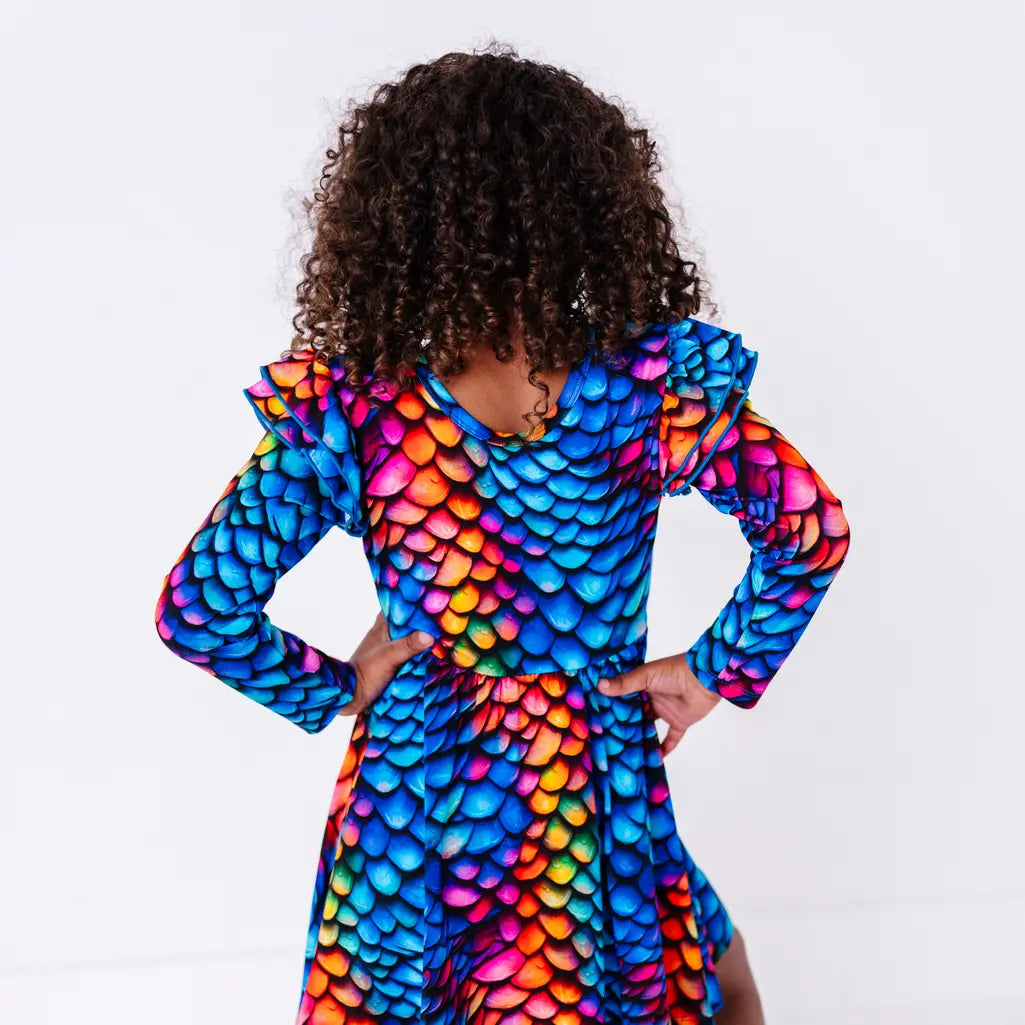 Back view of a child with curly hair and hands on the hips, in a long sleeve twirl dress with flutter ruffles at the shoulders in a rainbow dragon scale print of blue, pink, orange, and yellow.