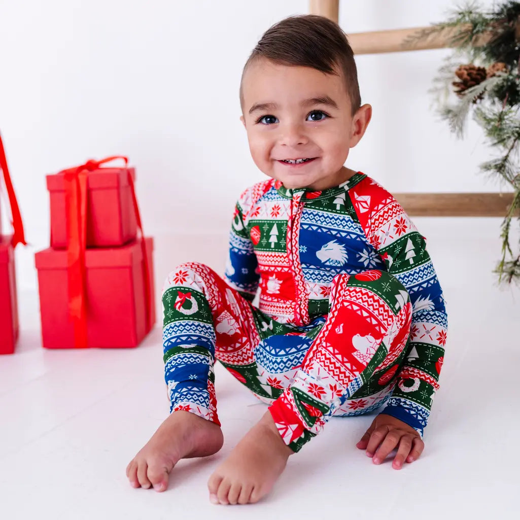 A smiling toddler sitting with knees up and bare feet, wearing a zip up baby pajama in red, green, blue, and white fair isle stripes with snowflakes, pine trees, and white dragon heads.
