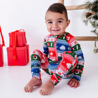 A smiling toddler sitting with knees up and bare feet, wearing a zip up baby pajama in red, green, blue, and white fair isle stripes with snowflakes, pine trees, and white dragon heads.