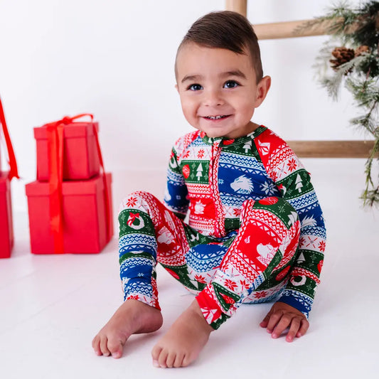 A smiling toddler sitting with knees up and bare feet, wearing a zip up baby pajama in red, green, blue, and white fair isle stripes with snowflakes, pine trees, and white dragon heads.